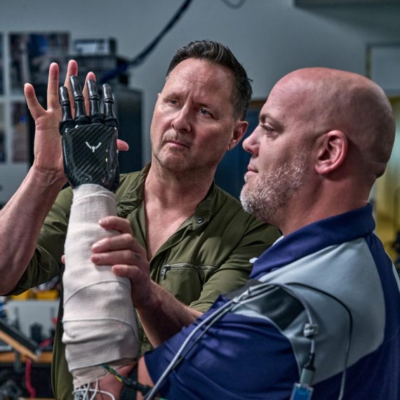 One man holds his hand up to the prosthetic hand of another man in a lab setting.