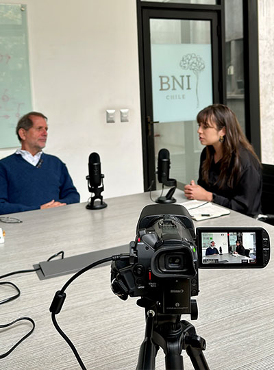 Woman interviews a man at a table with a camera recording the interview in the foreground.