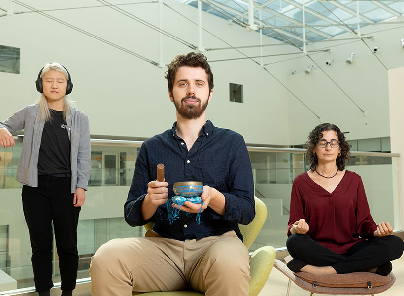 Three people practicing mindfulness in MIT Building 46. Woman on left is leaning on a railing, wearing headphones with eyes closed. Man seated in the center holds a bowl and a wooden spoon. Woman on right is seated with legs crossed and eyes closed. 