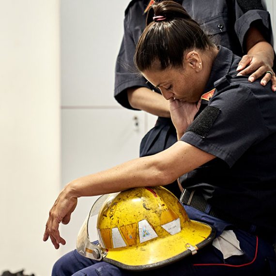 Side view of woman in uniform sitting on bench in fire station locker room with helmet in lap, head down, and eyes closed.