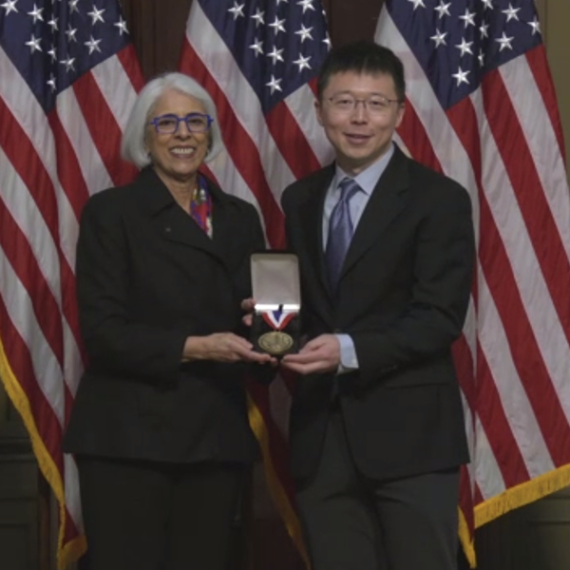 TWo people standing in front of American flags both holding a box with a medal in it.