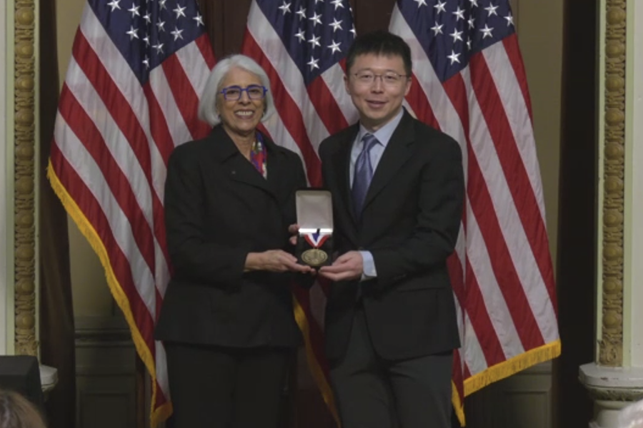 TWo people standing in front of American flags both holding a box with a medal in it.