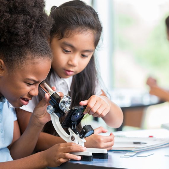 Focused African American and Asian schoolgirls help one another with their assignment in science class at a STEM elementary school. The African American girl is looking at something with a microscope.