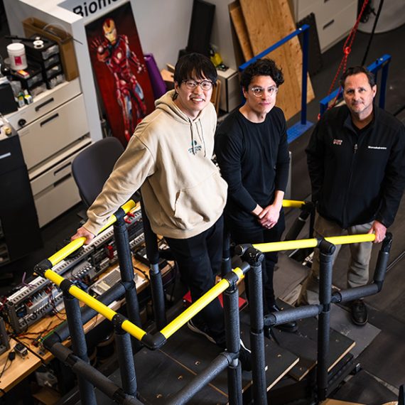 Three men stand on a makeshift staircase in a cluttered lab setting.