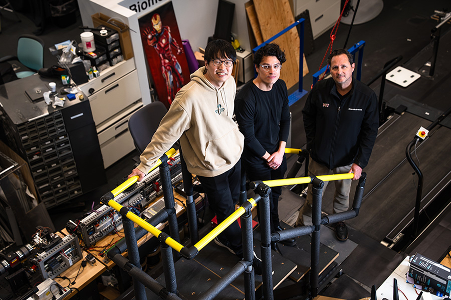 Three men stand on a makeshift staircase in a cluttered lab setting.