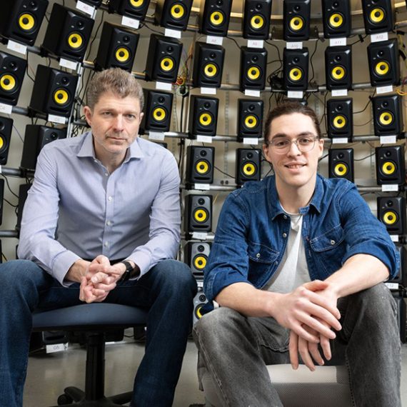 Two men seated in chairs, hands folded, in front of a curved wall of speakers.