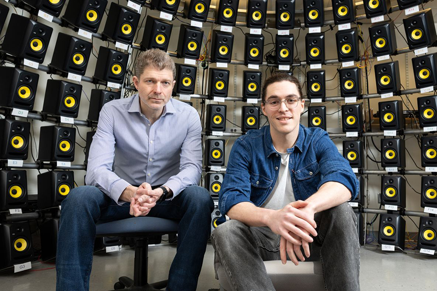Two men seated in chairs, hands folded, in front of a curved wall of speakers.